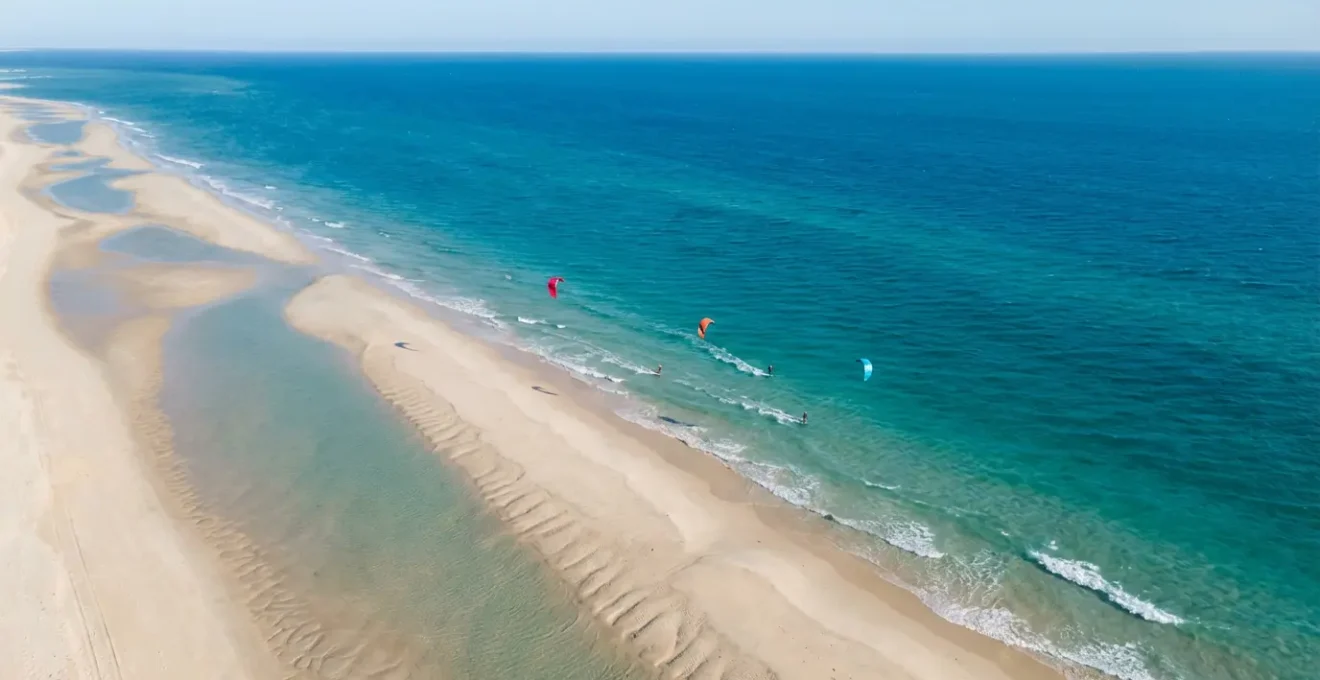 Vue aérienne grand angle d'une plage avec des flèches de vent montrant l'orientation side-shore idéale, kitesurfeurs évoluant parallèlement au rivage